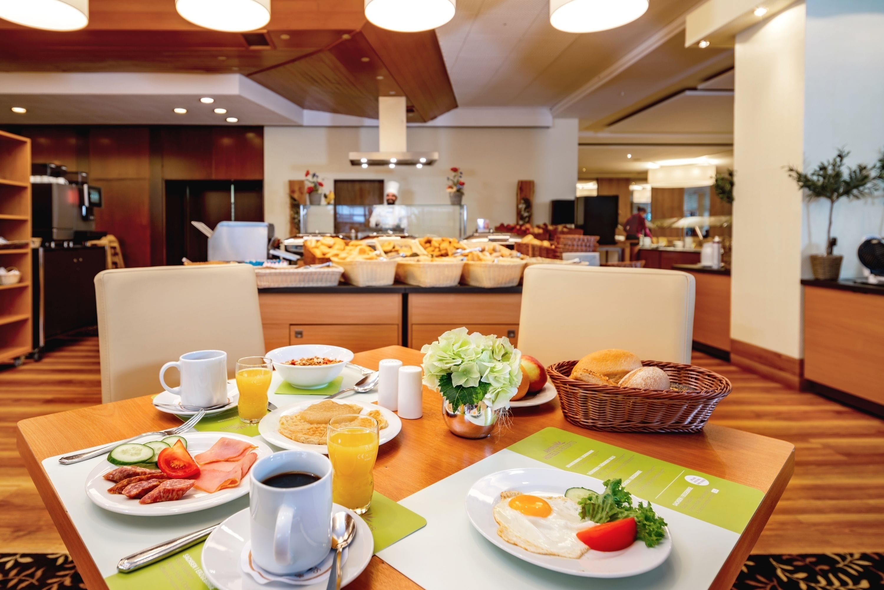 Um buffet de café da manhã com diversos alimentos e bebidas sobre uma mesa, uma cozinha ao fundo e uma área de refeições com cadeiras e mesa.