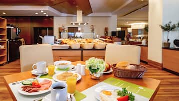 Um buffet de café da manhã com diversos alimentos e bebidas sobre uma mesa, uma cozinha ao fundo e uma área de refeições com cadeiras e mesa.