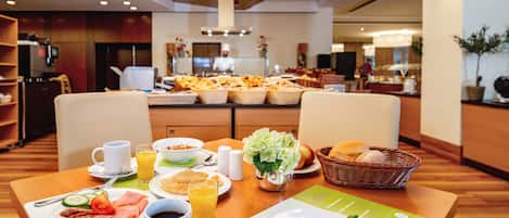 Un buffet de petit-déjeuner avec divers aliments et boissons disposés sur une table, un coin cuisine en arrière-plan et un espace repas avec des chaises et une table.