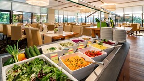 Een buffet met diverse gerechten, waaronder salades, granen en groenten, in een goed verlichte eetruimte met grote ramen en modern meubilair.