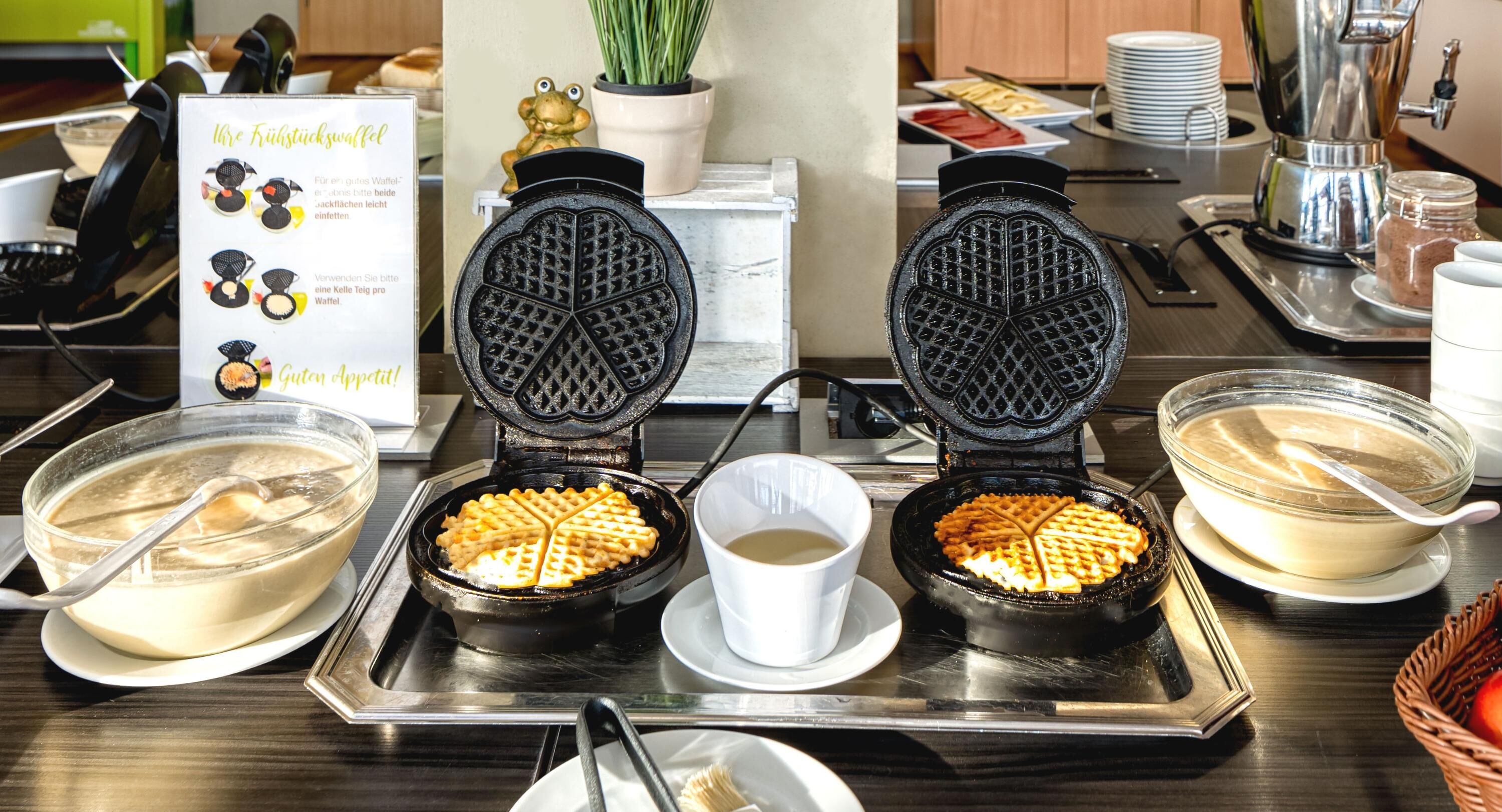 Ein Waffelstand mit zwei Waffeleisen, einer Schüssel Teig, einer Kaffeetasse und einem Marmeladenglas.