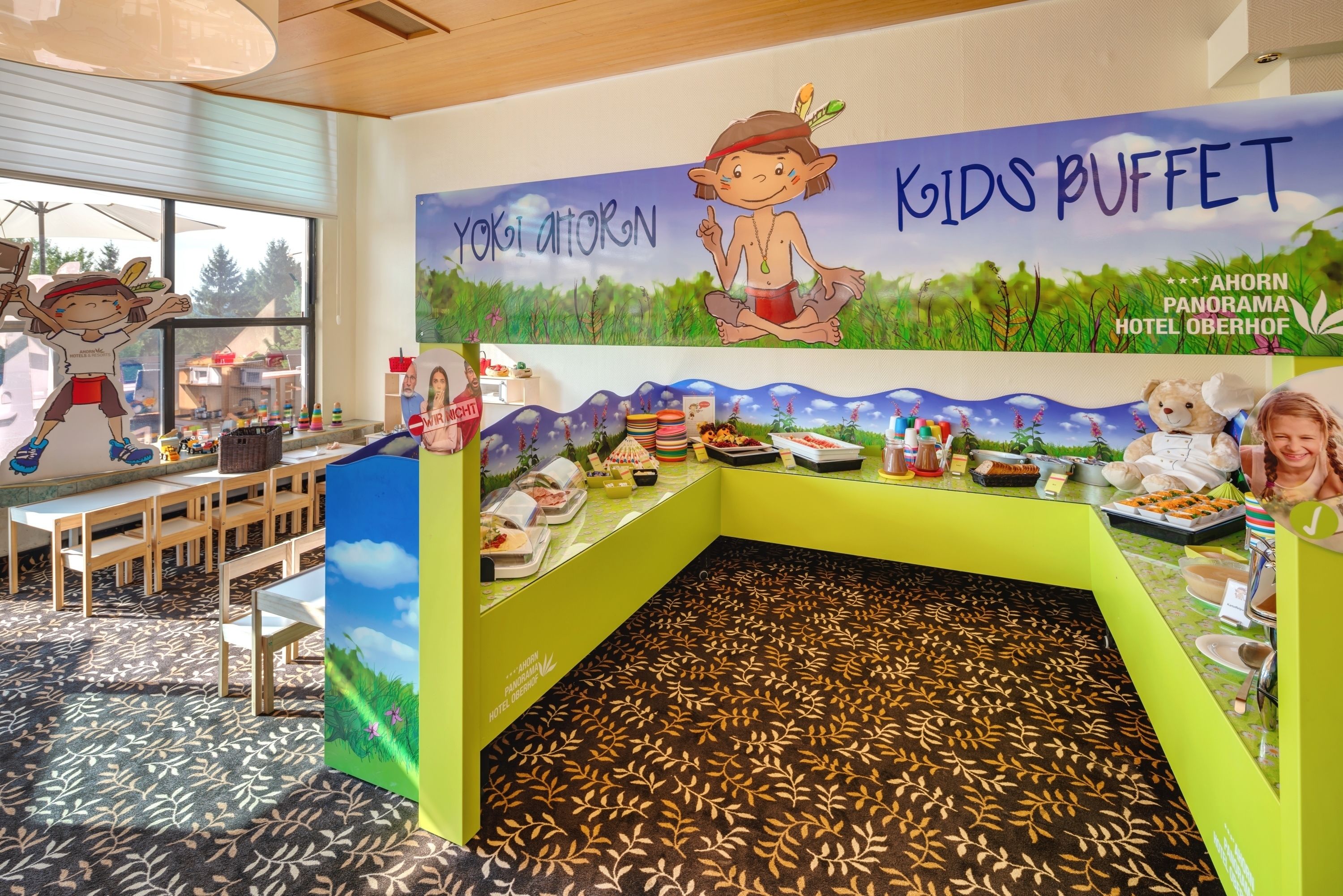 A children's buffet area with a mural of a child and nature, tables with food, and a patterned carpet.