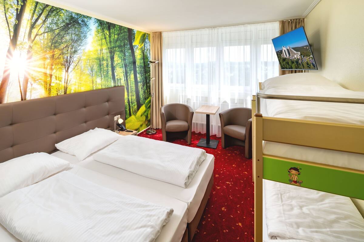 A hotel room with a large bed, a smaller bed, a desk, two chairs, and a wall-mounted TV.
