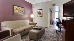 A hotel room with a beige sofa, a small ottoman, a TV, a lamp, and a patterned carpet. - Drury Inn & Suites San Antonio Northeast (San Antonio)
