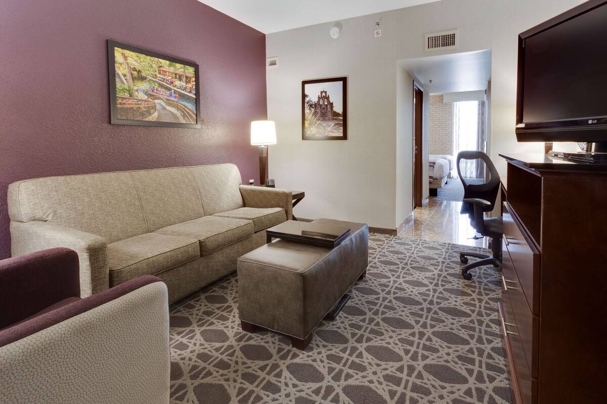 A hotel room with a beige sofa, a small ottoman, a TV, a lamp, and a patterned carpet.