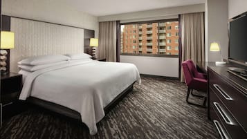 A hotel room with a large bed, a desk with a TV, a chair, and a view of a building through the window.