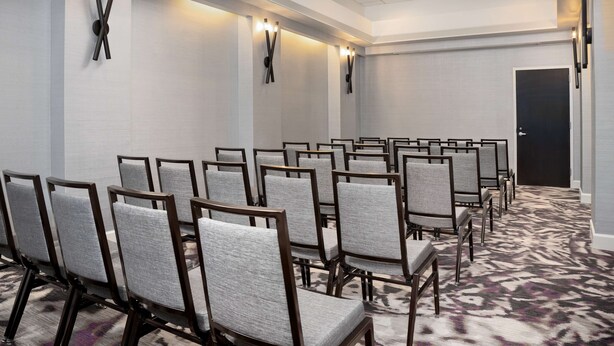 Un salón de conferencias con filas de sillas tapizadas en gris, alfombra estampada y luminarias fijadas a la pared.