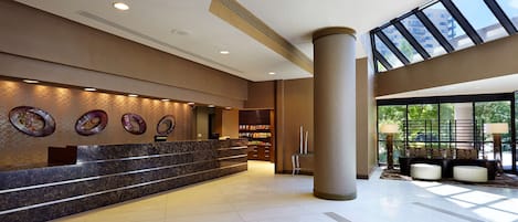 A modern hotel lobby with a marble reception desk, decorative wall art, a seating area with a view of the outdoors, and a large window.