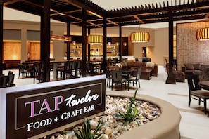 Hyvin valaistu sisätila, jossa on kyltti, jossa lukee ”Tap Twelve Food + Craft Bar”. Tilassa on pöytiä ja tuoleja, baarialue sekä koristeellinen kasviasetelma keskellä.