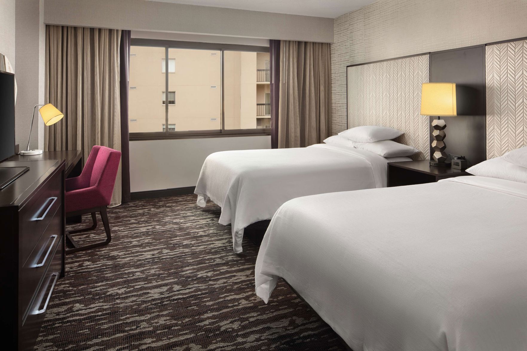 A hotel room with two beds, a desk, a chair, and a large window with curtains.