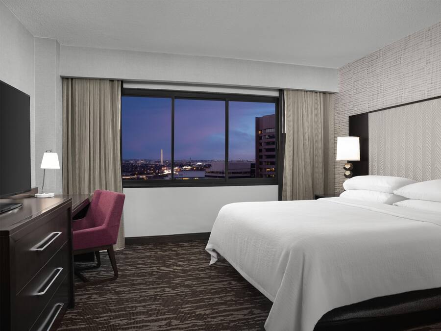 Quarto de hotel com cama, escrivaninha, cadeira, televisão e janela com vista para a cidade.