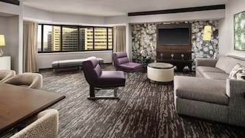 A hotel room with a large window, a flat-screen TV, a sofa, a chair, a coffee table, and a carpeted floor.