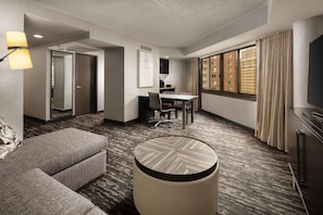 Habitación de hotel con sofá, una mesa redonda, escritorio con silla, televisor y ventana con cortinas.