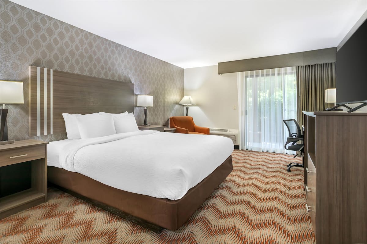 Habitación de hotel con una cama grande, dos mesitas de noche con lámparas, una silla naranja, un escritorio con una silla y un ventanal con cortinas.