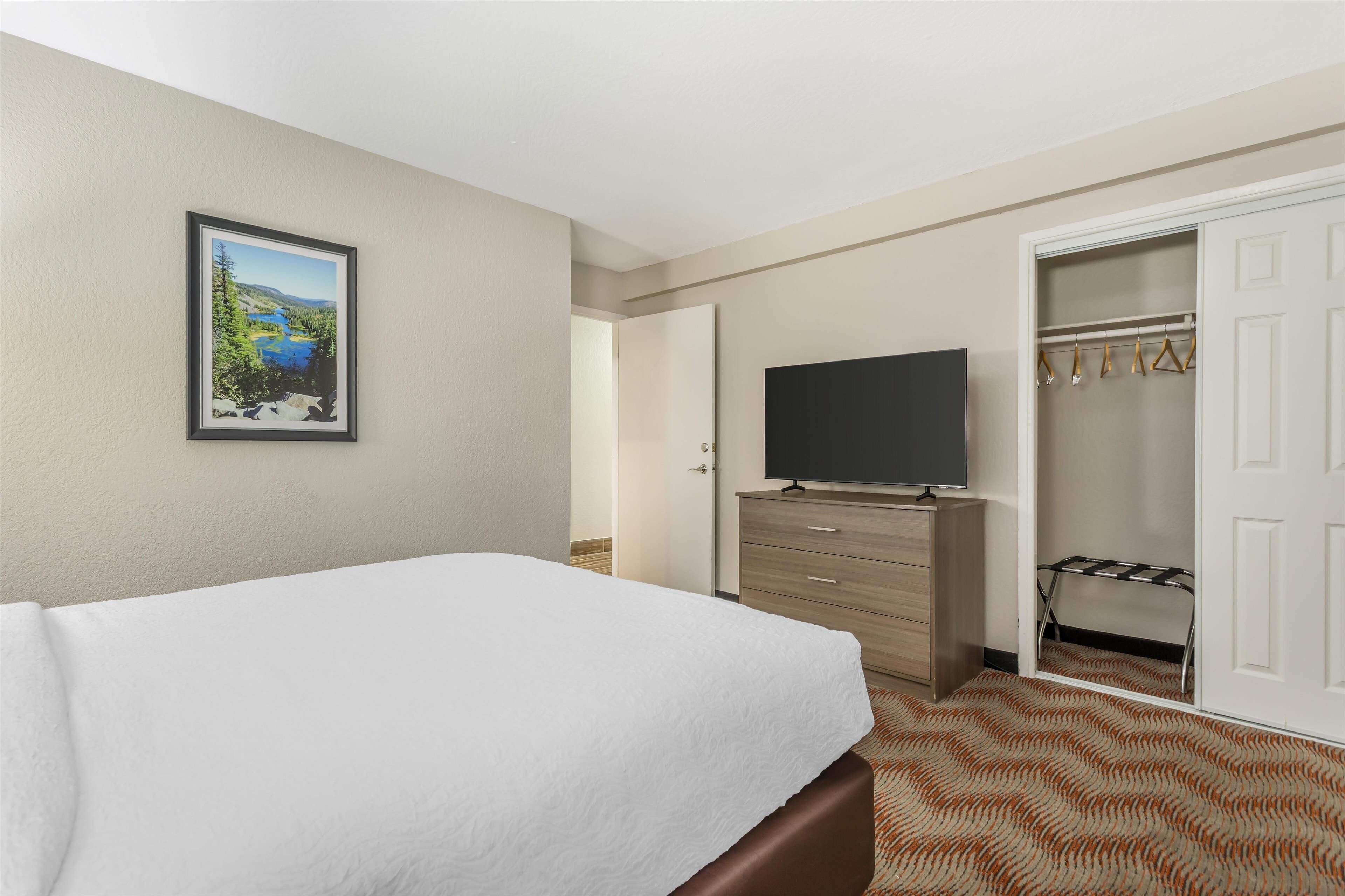 A hotel room with a bed, a television on a dresser, a closet with hangers, and a door leading to another room.