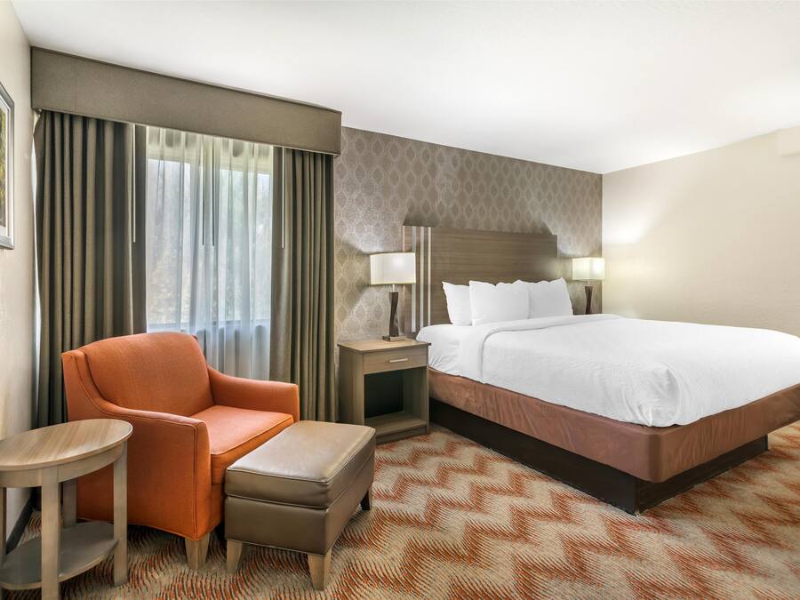 A hotel room with a large bed, a chair, a small table, and a nightstand with lamps.