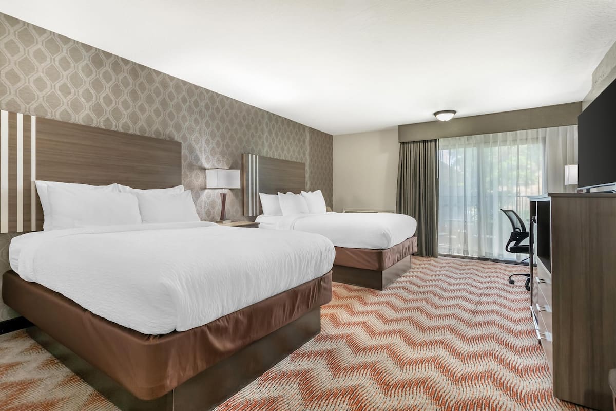 Una habitación de hotel con dos camas, un escritorio, una lámpara y un ventanal grande con cortinas.