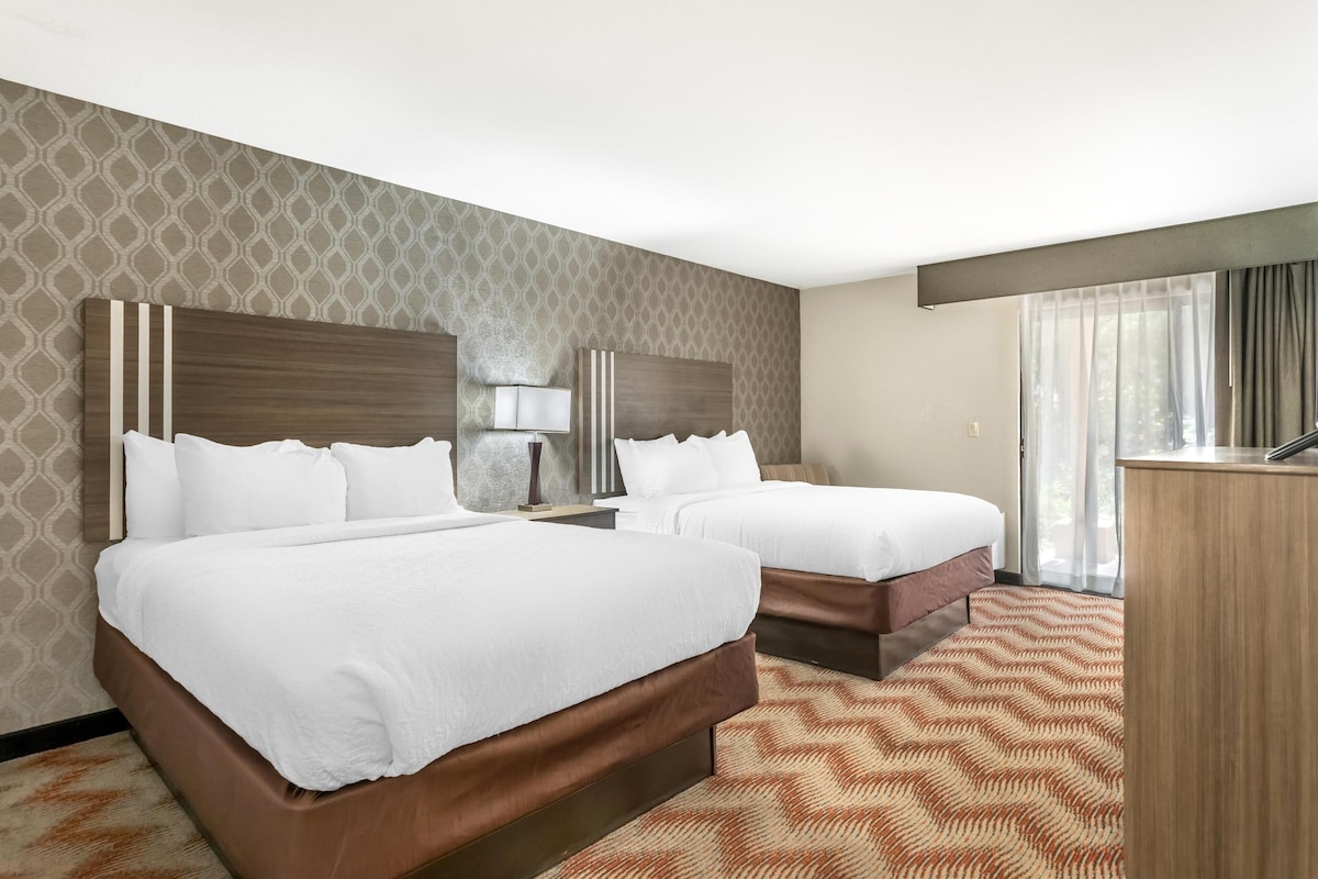 A hotel room with two beds, a desk, and a large window with curtains.