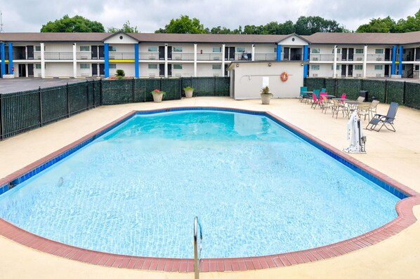 A swimming pool with a brick border, surrounded by a fence, and a building with balconies in the background.