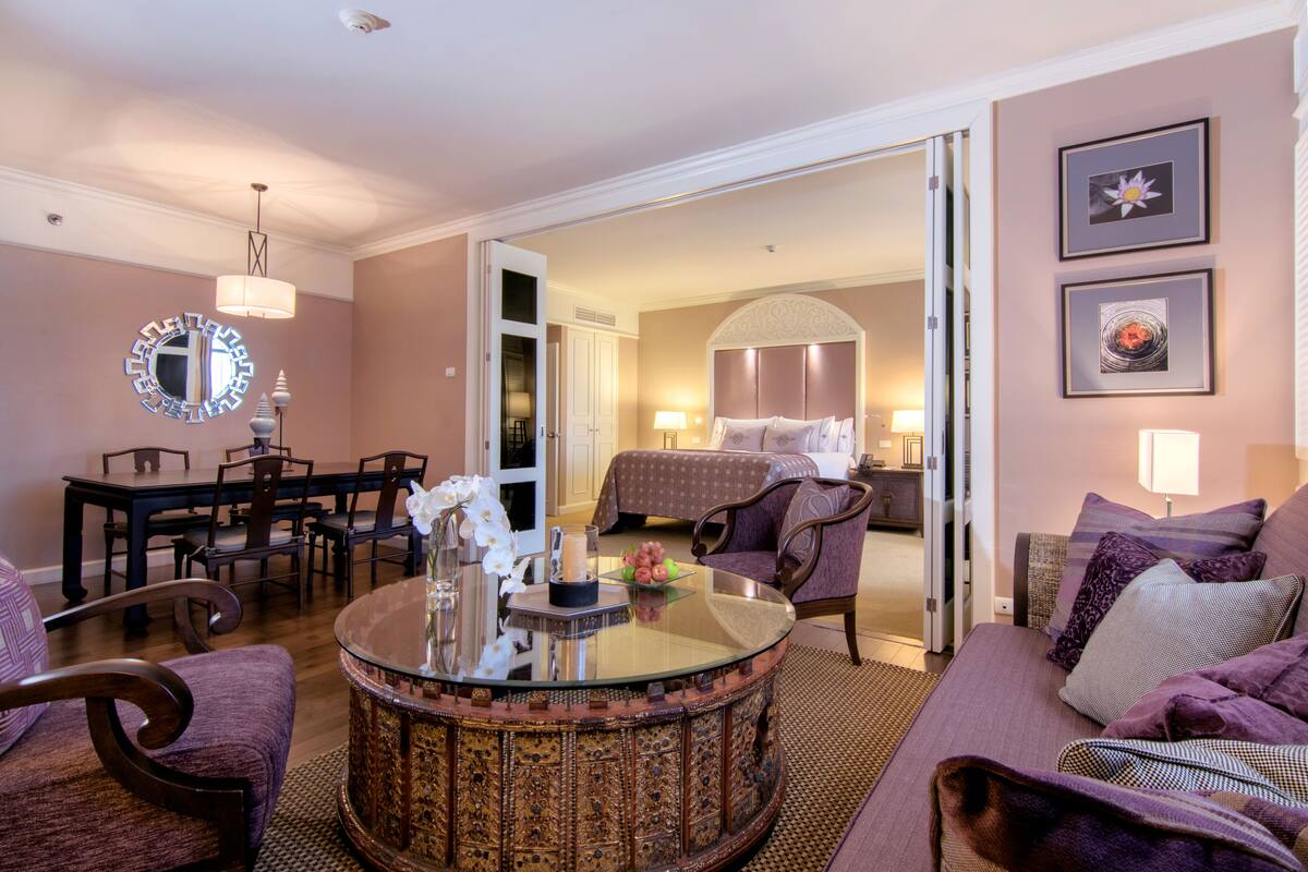 A hotel room with a round glass coffee table, a sofa, a dining area with chairs, and a bed with a headboard.