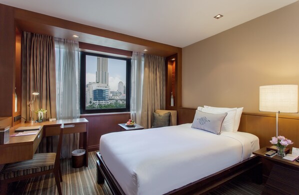 Habitación de hotel con una cama grande, un escritorio, una silla, una mesita con un jarrón de flores y vistas a la silueta urbana a través de un ventanal amplio.