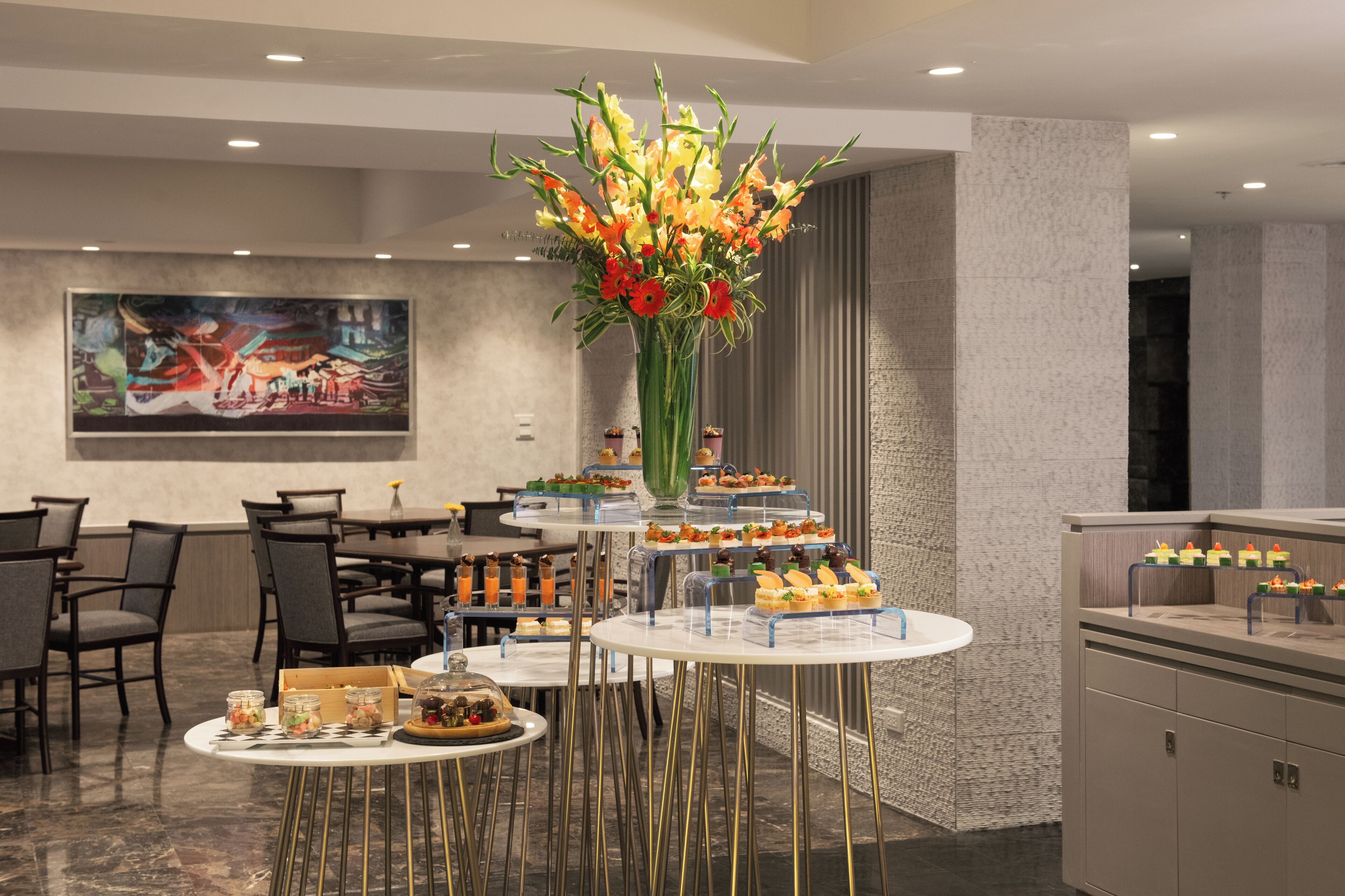 A modern dining area with round tables, a tiered dessert display, and a large floral centerpiece.