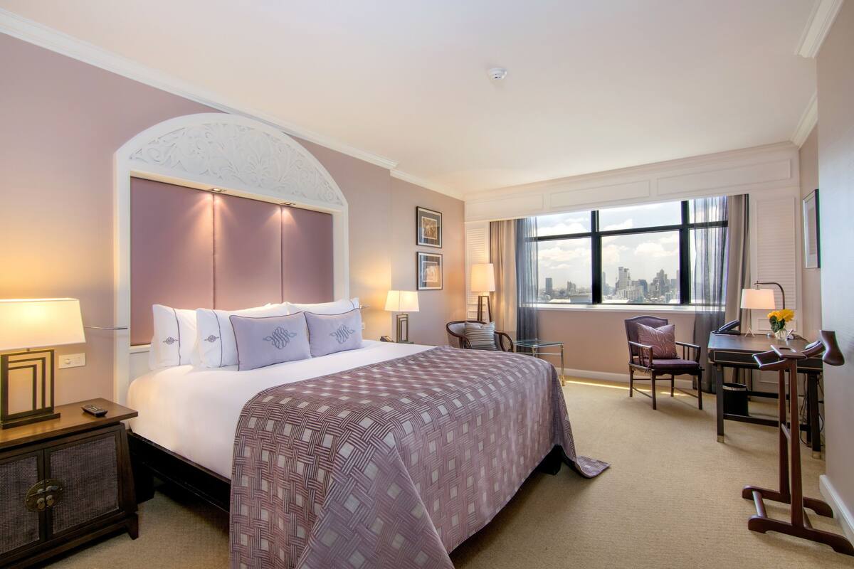 A hotel room with a large bed, a desk, a chair, and a view of the city through the window.