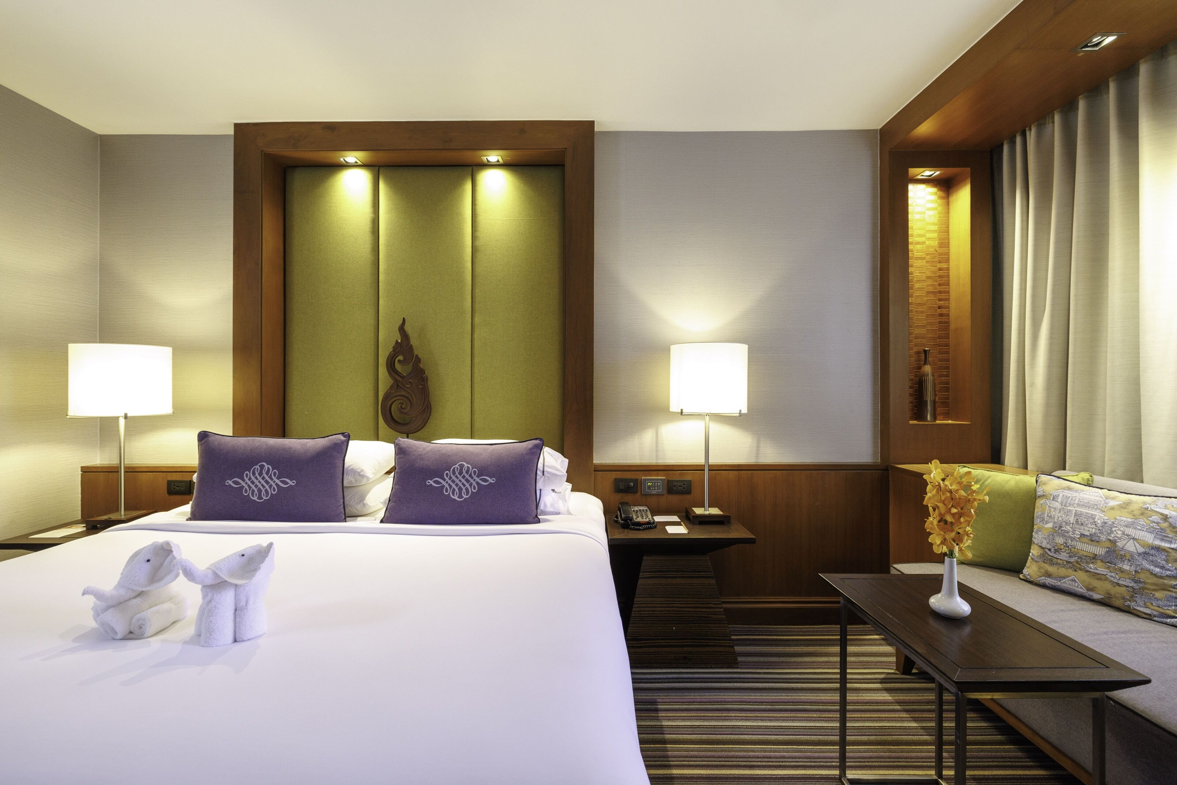 A hotel room with a bed, two bedside tables, a nightstand, a sofa, and a small table with a vase of flowers.