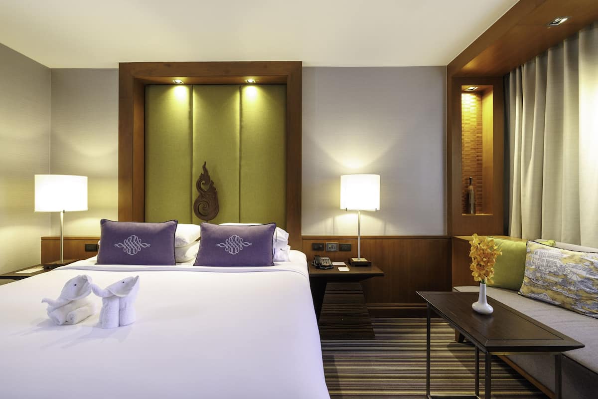 A hotel room with a bed, two bedside tables, a nightstand, a sofa, and a small table with a vase of flowers.