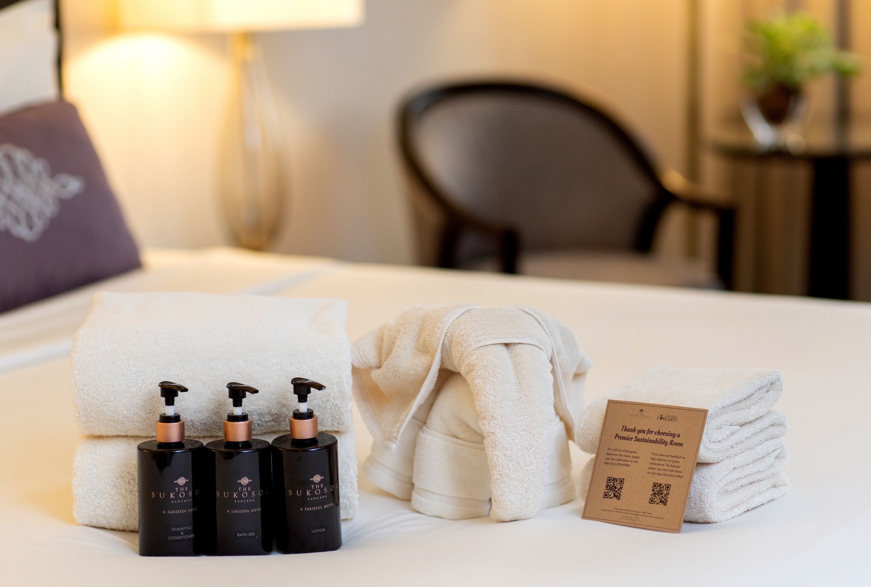 A neatly made bed with white towels, a folded towel, and three bottles of shampoo and bath oil. A thank you card for choosing a sustainable room is also visible.