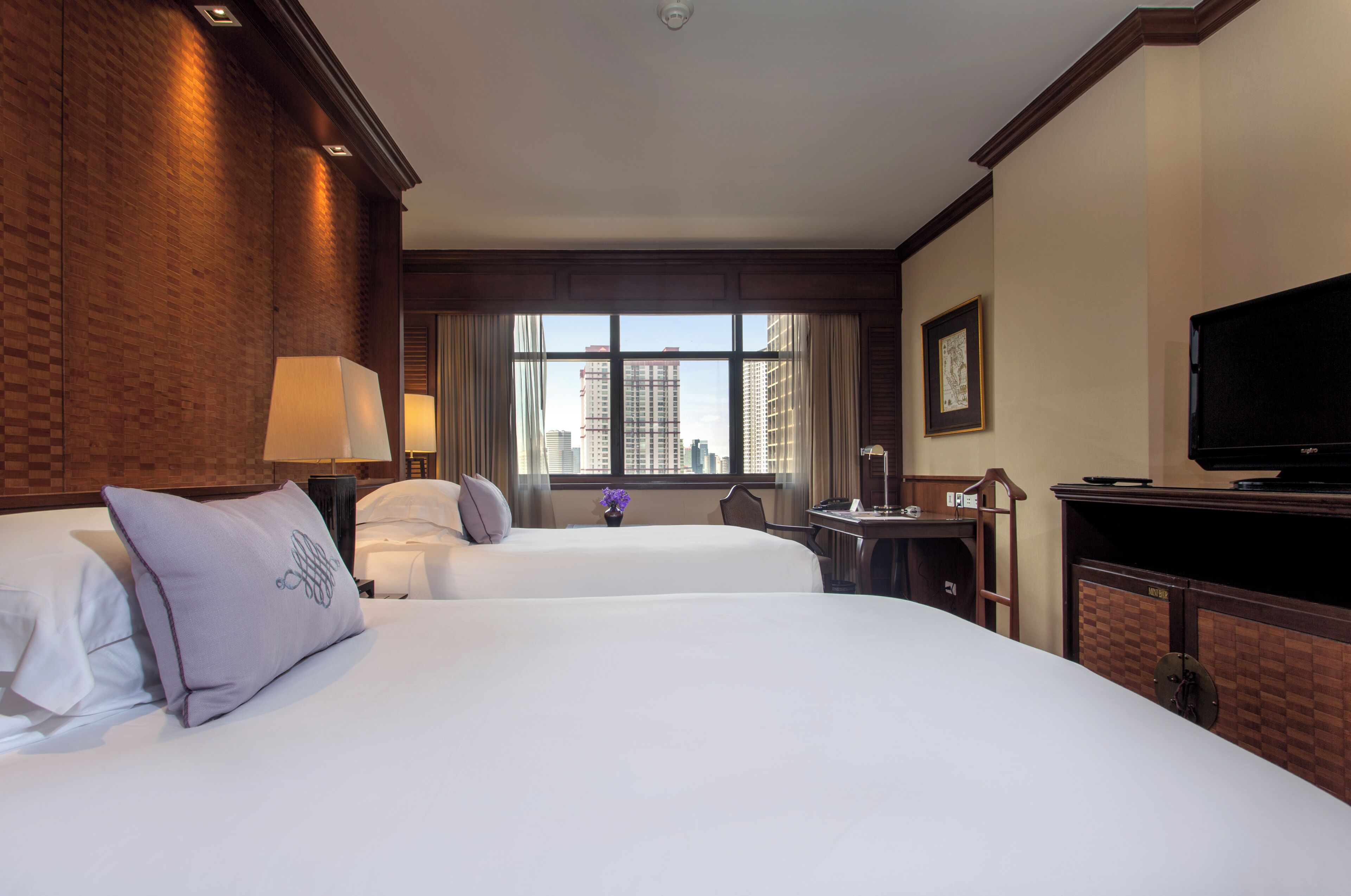 A hotel room with two beds, a desk, a television, and a window with a city view.