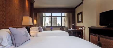A hotel room with two beds, a desk, a television, and a window with a city view.