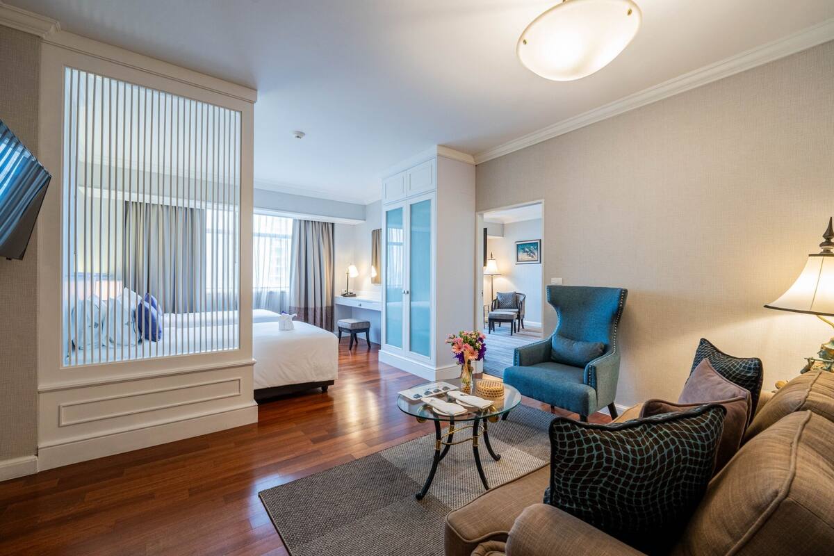A hotel room with a bed, a sitting area with a sofa and armchairs, a small table with a vase, and a large window with blinds.