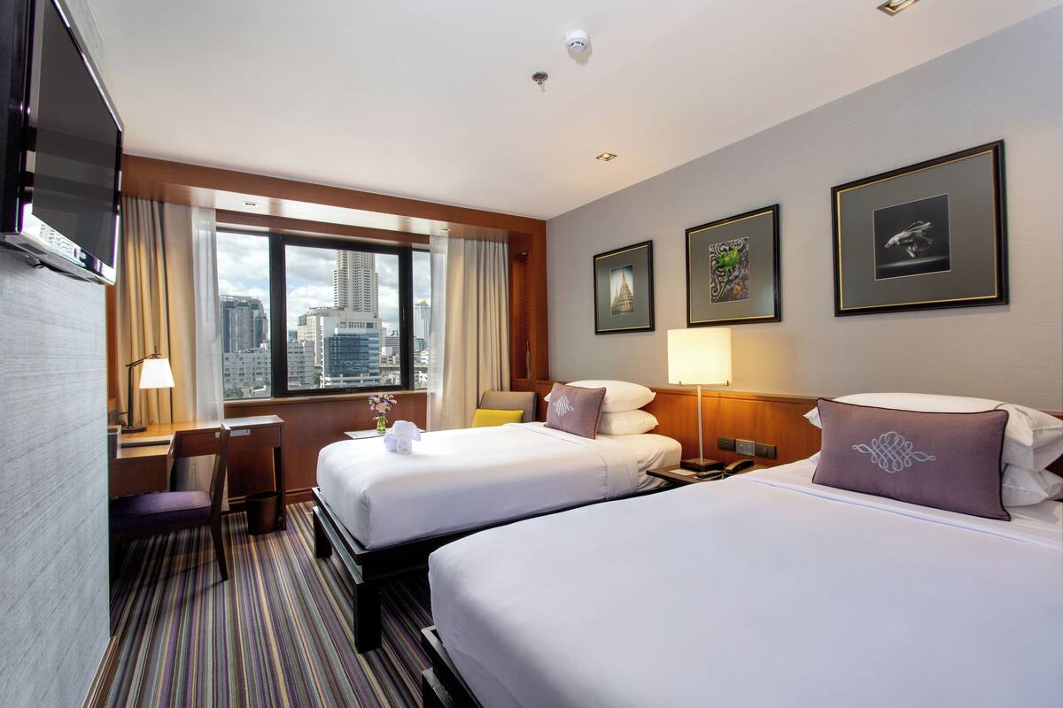 A hotel room with two beds, a desk, a chair, a TV, a window with a city view, and framed artwork on the wall.