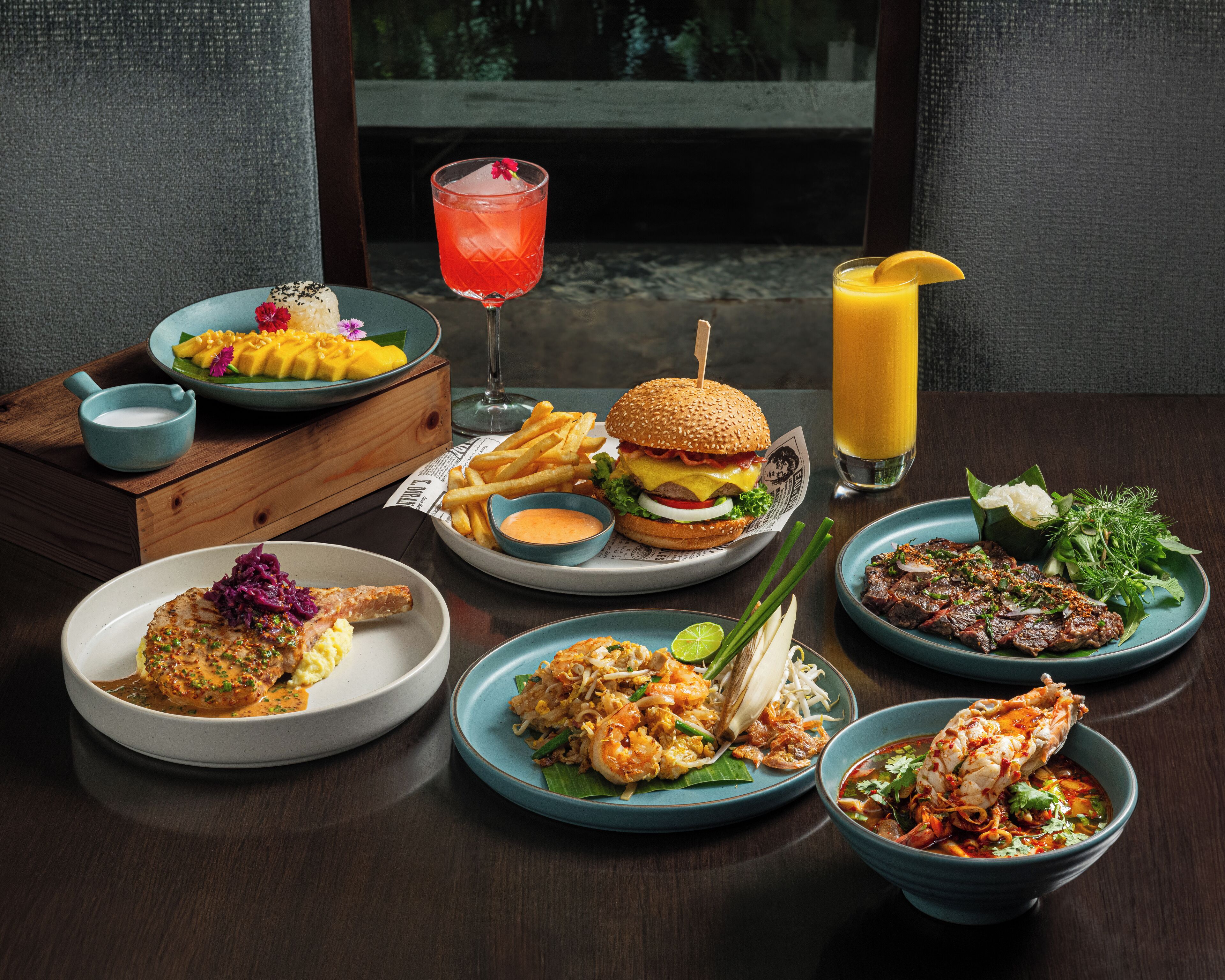 A variety of dishes including a burger, seafood, and a salad, arranged on a wooden table with a view of a window in the background.