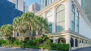 A building with large arched windows and a white facade, surrounded by lush greenery and palm trees. - The Sukosol (Bangkok)