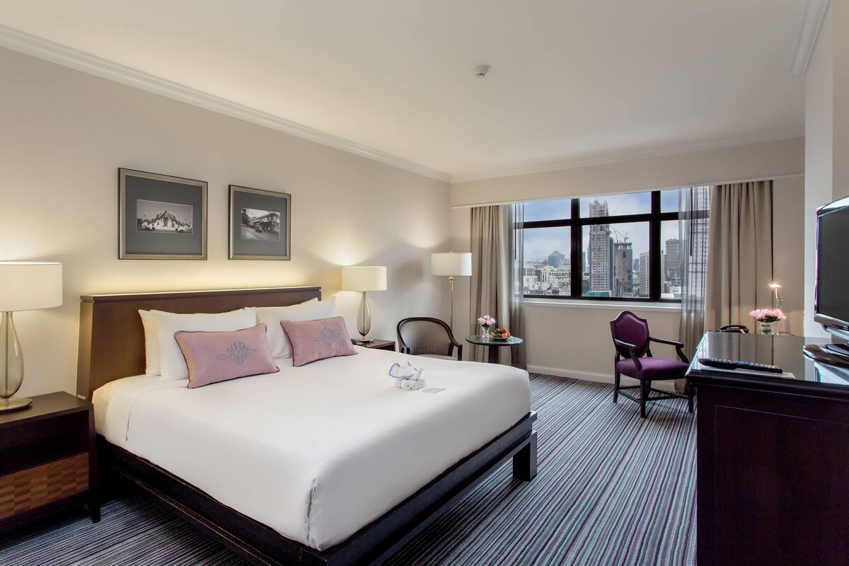 A hotel room with a large bed, two bedside tables with lamps, a desk with a chair, a television, and a window with a city view.