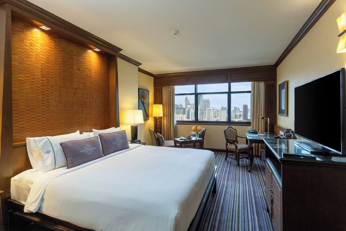 A hotel room with a large bed, a desk, a chair, a TV, and a window with a city view.
