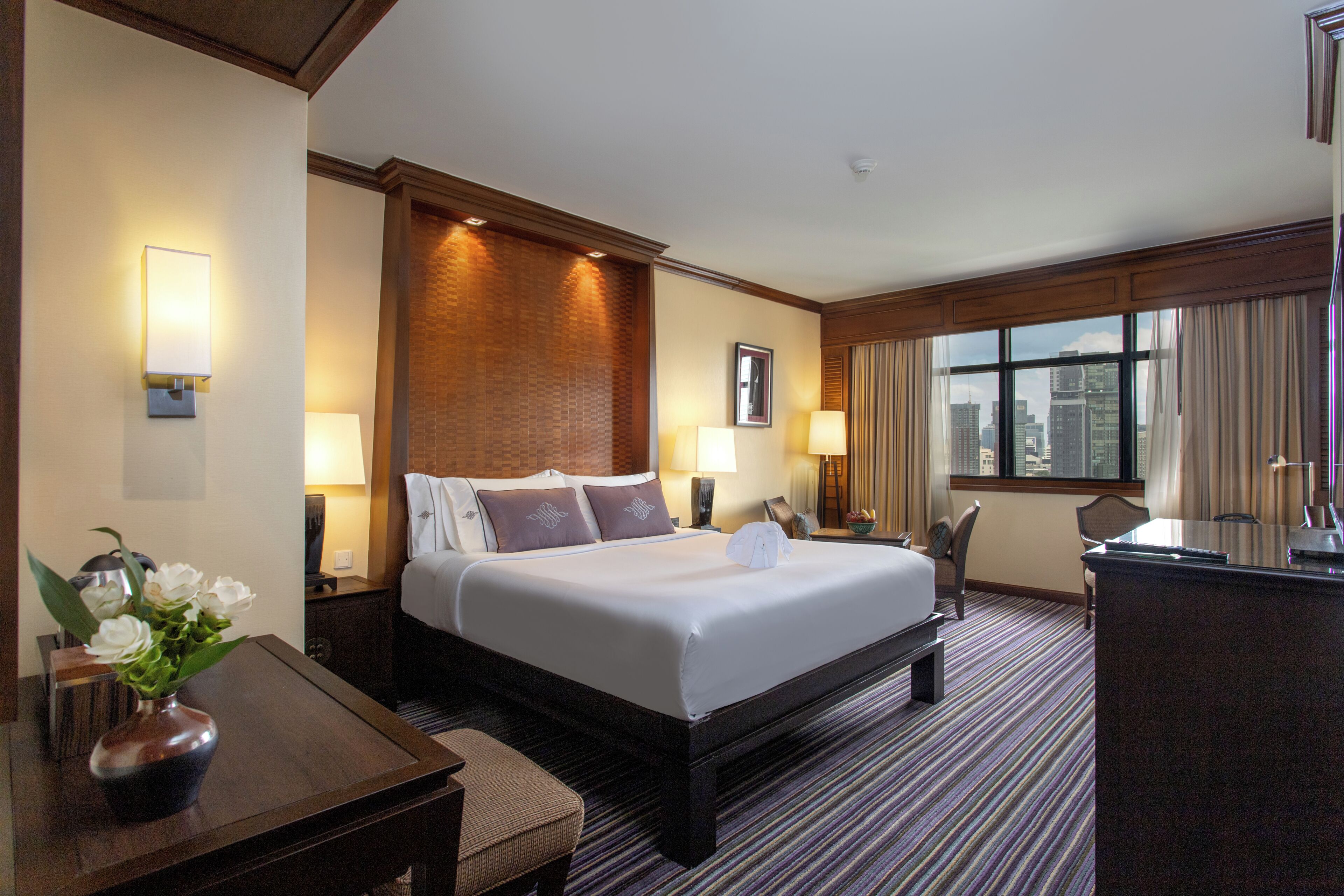 A hotel room with a large bed, bedside lamps, a desk, and a view of the city through the window.