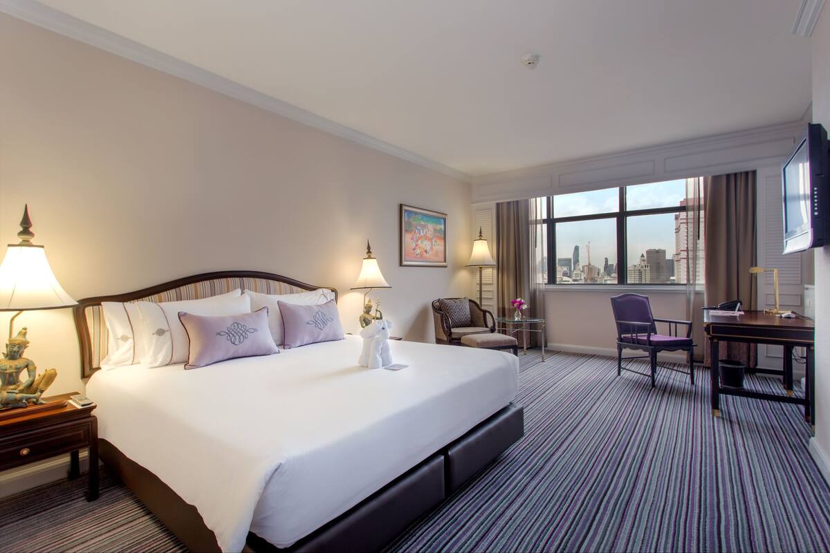 A hotel room with a large bed, two bedside lamps, a desk with a chair, a TV, and a window with a city view.