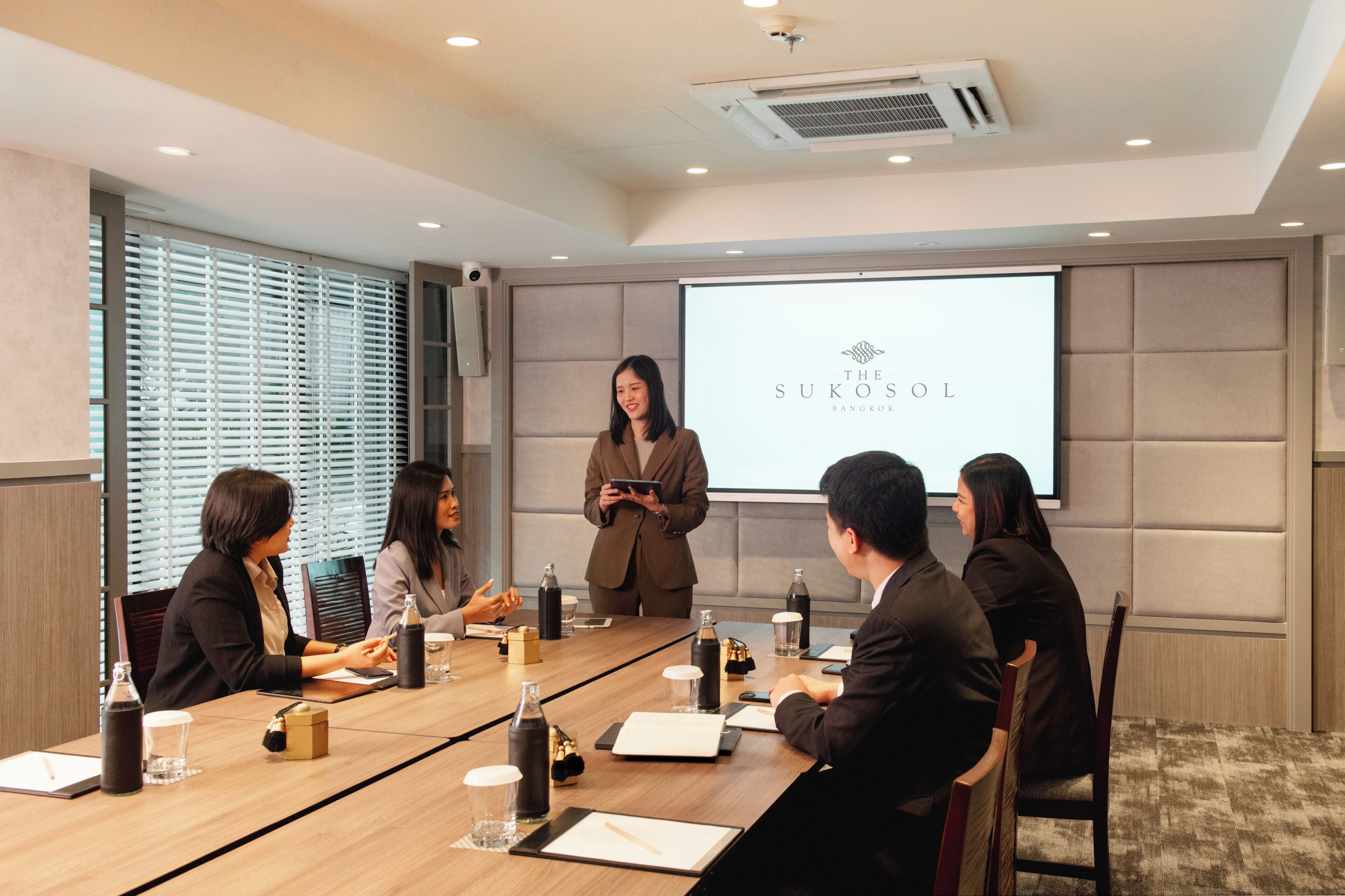 Un salón de conferencias con una pantalla mostrando el logotipo de 'The Sukosol Bangkok', un grupo de personas sentadas alrededor de una mesa con computadoras portátiles, y una persona presentando de pie.