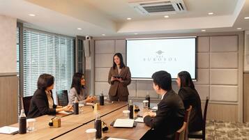 Un salón de conferencias con una pantalla mostrando el logotipo de 'The Sukosol Bangkok', un grupo de personas sentadas alrededor de una mesa con computadoras portátiles, y una persona presentando de pie.