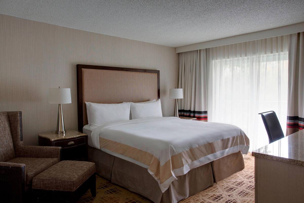 A hotel room with a large bed, a nightstand with a lamp, a chair, and a desk.