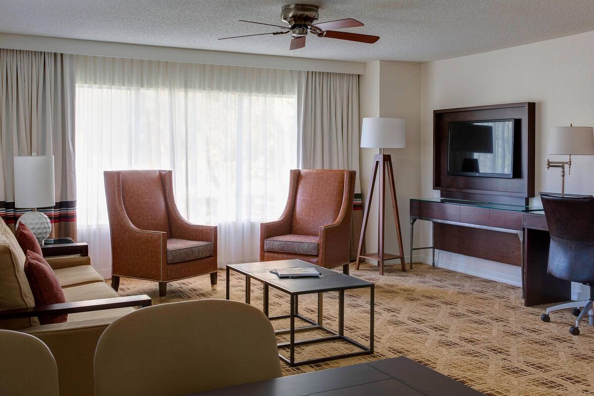 A hotel room with two leather armchairs, a sofa, a small coffee table, a desk with a flat-screen TV, a lamp, and a ceiling fan.