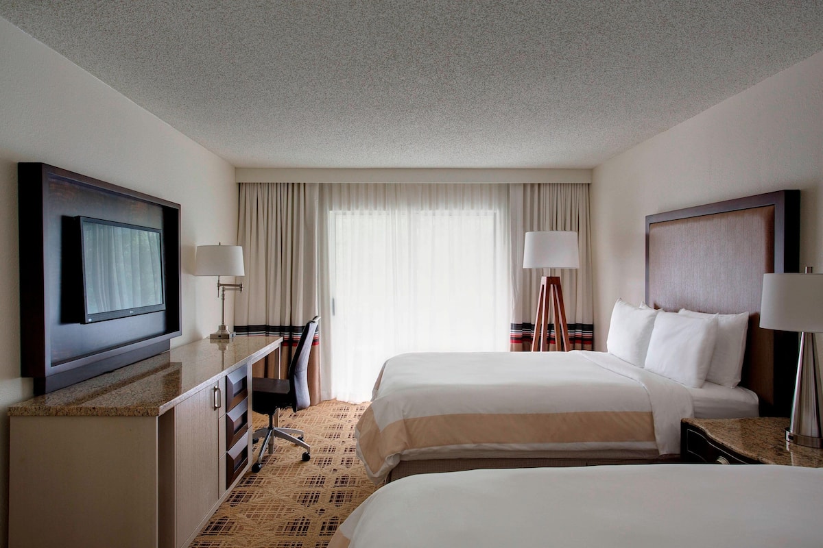 A hotel room with two beds, a desk with a TV, and a large window with curtains.