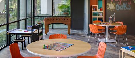Una sala de juegos infantil moderna con ventanas amplias, sillas naranjas, una mesa de juegos de madera y una pared con pizarrón.