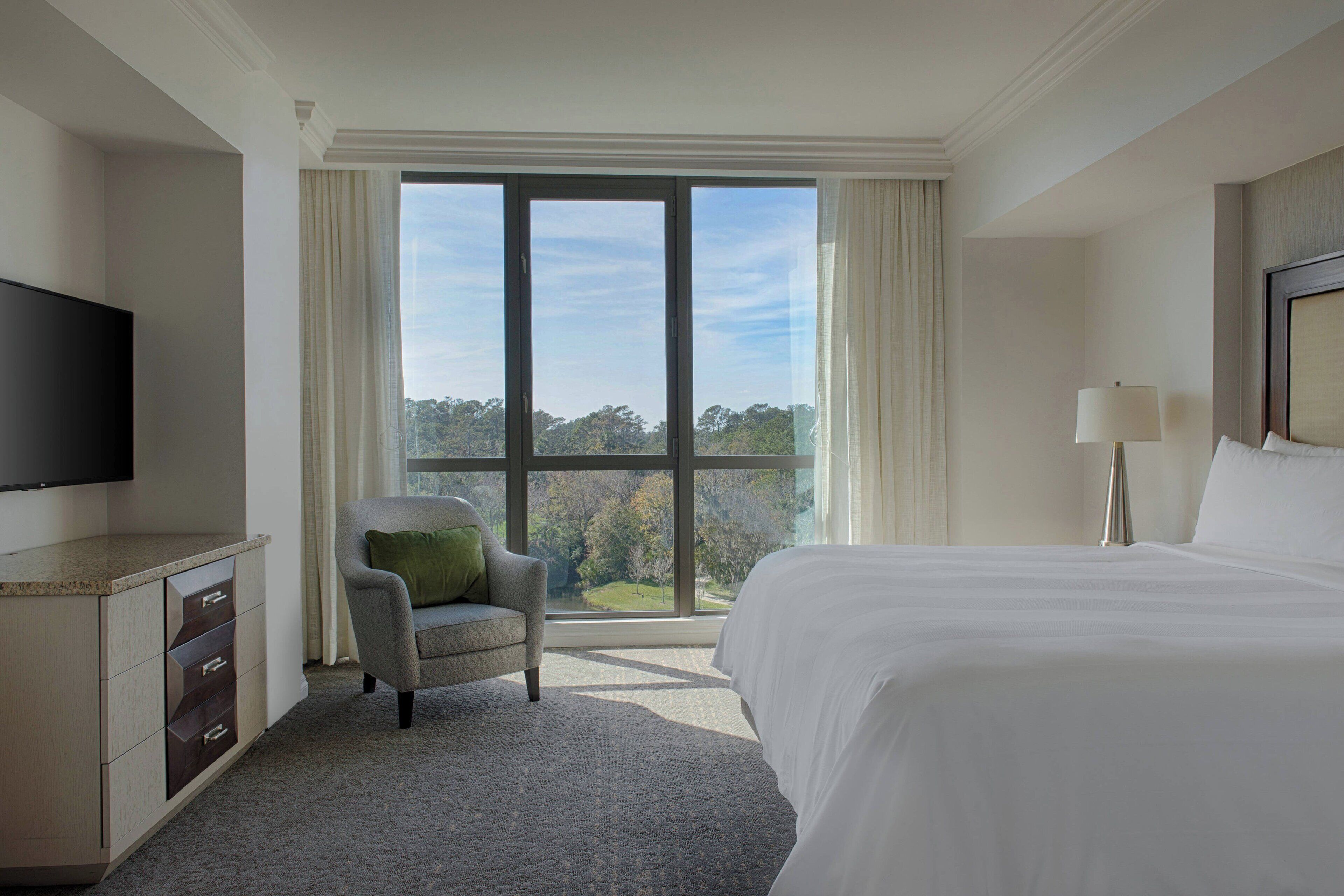 A hotel room with a large bed, a flat-screen TV, a chair, and a view of the outdoors through a large window.