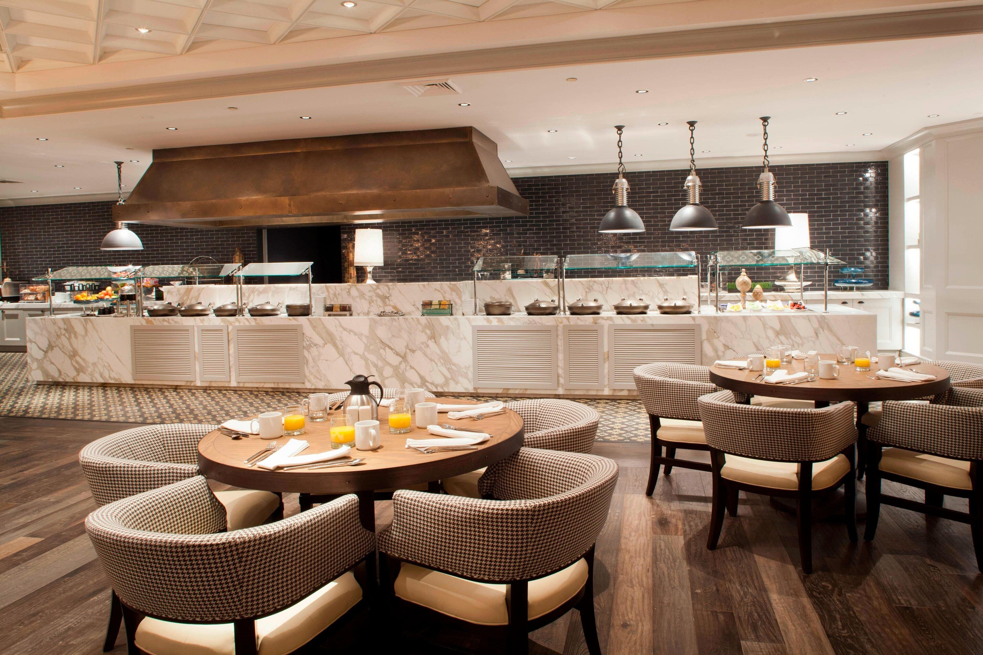 A modern restaurant with round tables and checkered chairs, a marble bar, and a large kitchen in the background.