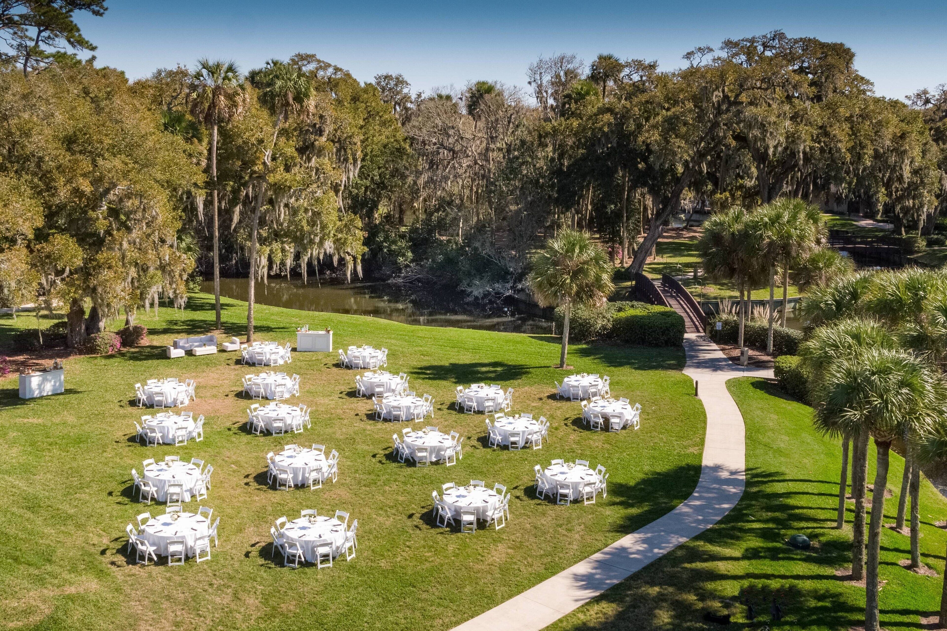 A well-maintained outdoor area with multiple round tables and chairs arranged on a grassy lawn, surrounded by trees and a pathway leading to a building.