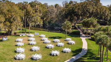 A well-maintained outdoor area with multiple round tables and chairs arranged on a grassy lawn, surrounded by trees and a pathway leading to a building.